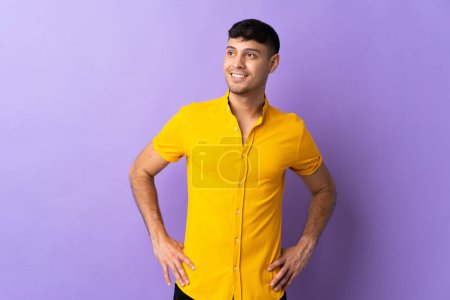 Photo for Young Colombian man isolated on purple background posing with arms at hip and smiling - Royalty Free Image