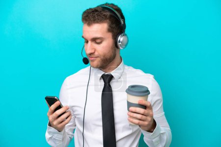 Téléchargez les photos : Telemarketer caucasian man working with a headset isolated on blue background holding coffee to take away and a mobile - en image libre de droit