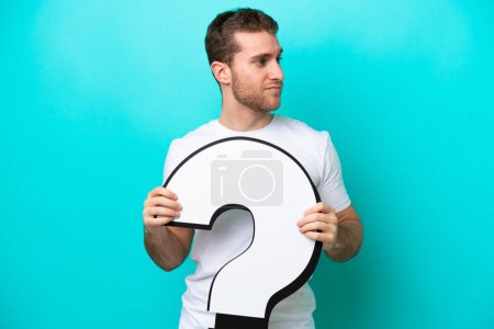 Photo for Young caucasian man isolated on blue background holding a question mark icon and looking side - Royalty Free Image