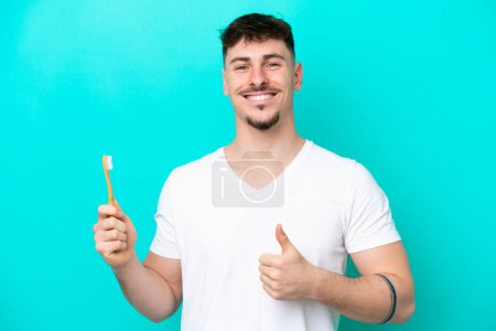Téléchargez les photos : Jeune homme caucasien brossant les dents isolé sur fond bleu avec les pouces vers le haut parce que quelque chose de bon est arrivé - en image libre de droit