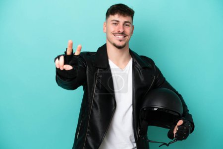 Foto de Joven hombre caucásico con un casco de moto aislado sobre fondo azul sonriendo y mostrando señal de victoria - Imagen libre de derechos
