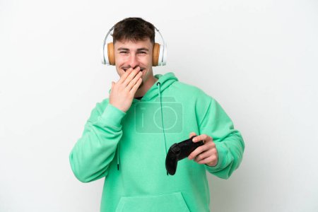 Foto de Joven hombre guapo jugando con un controlador de videojuego aislado sobre fondo blanco feliz y sonriente boca cubierta con la mano - Imagen libre de derechos