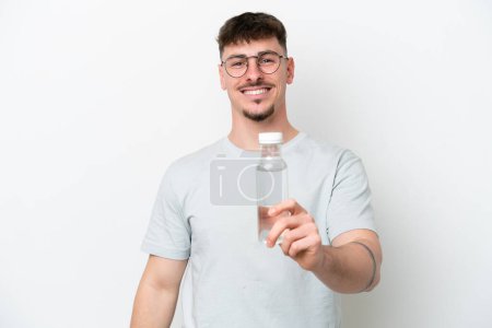 Téléchargez les photos : Jeune homme caucasien tenant une bouteille d'eau isolée sur fond blanc avec une expression heureuse - en image libre de droit
