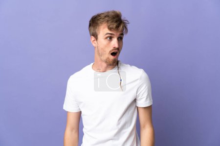 Photo for Young handsome blonde man isolated on purple background doing surprise gesture while looking to the side - Royalty Free Image
