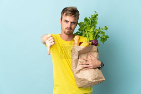 Photo for Blonde Person taking a bag of takeaway food isolated on blue background showing thumb down with negative expression - Royalty Free Image