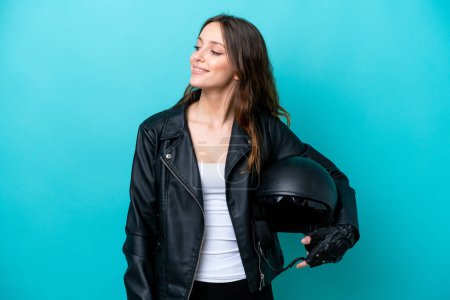 Téléchargez les photos : Jeune femme caucasienne avec un casque de moto isolé sur fond bleu regardant sur le côté et souriant - en image libre de droit