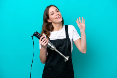 Téléchargez les photos : Jeune cuisinière caucasienne femme utilisant mélangeur à main isolé sur fond bleu saluant à la main avec expression heureuse - en image libre de droit