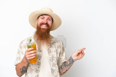 Téléchargez les photos : Rousse homme à longue barbe buvant un cocktail sur une plage isolée sur fond blanc pointant du doigt sur le côté - en image libre de droit