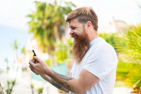 Foto de Pelirrojo con barba enviando un mensaje o correo electrónico con el móvil - Imagen libre de derechos