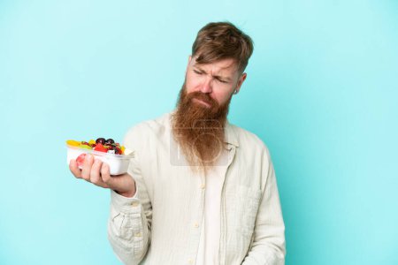 Téléchargez les photos : Homme rousse à longue barbe tenant un bol de fruits isolé sur fond bleu avec une expression triste - en image libre de droit