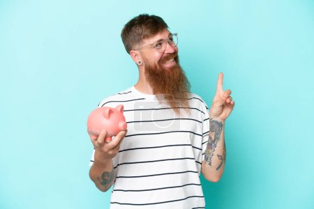 Téléchargez les photos : Homme rousse à longue barbe tenant une tirelire isolée sur fond bleu dans l'intention de réaliser la solution tout en levant le doigt - en image libre de droit