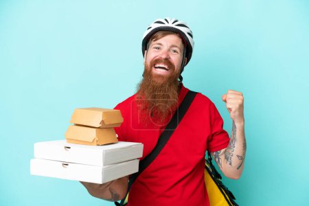 Foto de Repartidor sosteniendo pizzas y hamburguesas aisladas sobre fondo azul celebrando una victoria en posición ganadora - Imagen libre de derechos