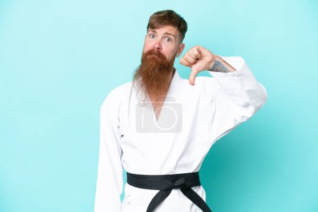 Foto de Hombre pelirrojo con barba larga haciendo karate aislado sobre fondo azul mostrando el pulgar hacia abajo con expresión negativa - Imagen libre de derechos