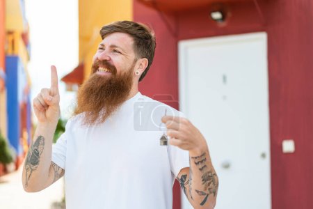 Téléchargez les photos : Homme rousse avec barbe tenant les clés de la maison à l'extérieur dans l'intention de réaliser la solution tout en levant le doigt - en image libre de droit