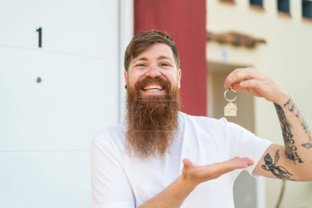 Téléchargez les photos : Homme rousse avec barbe tenant les clés de la maison avec une expression heureuse - en image libre de droit