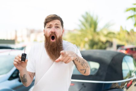 Téléchargez les photos : Homme rousse avec barbe tenant les clés de voiture à l'extérieur surpris et pointant devant - en image libre de droit