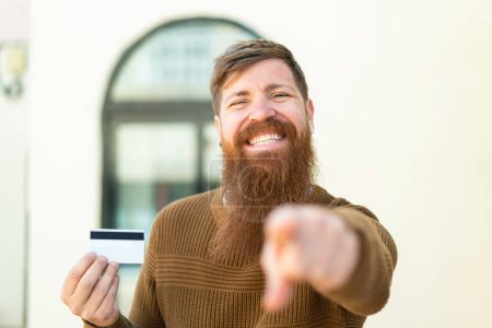Téléchargez les photos : Rousse homme avec barbe tenant une carte de crédit à l'extérieur pointe doigt à vous avec une expression confiante - en image libre de droit