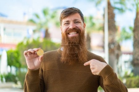 Foto de Pelirrojo con barba sosteniendo un Bitcoin al aire libre con expresión facial sorpresa - Imagen libre de derechos