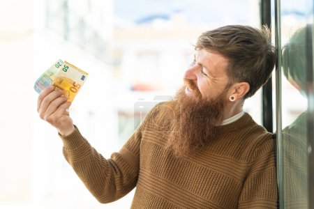 Téléchargez les photos : Rousse homme avec barbe prendre beaucoup d'argent à l'extérieur avec expression heureuse - en image libre de droit
