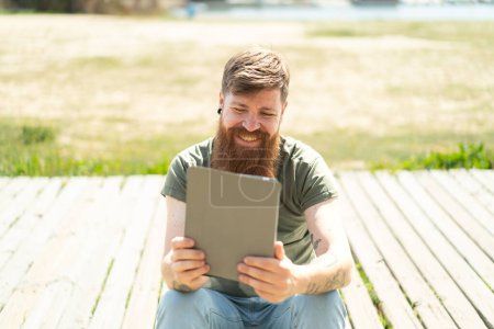Téléchargez les photos : Homme rousse avec barbe tenant une tablette avec une expression heureuse - en image libre de droit