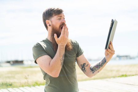 Téléchargez les photos : Homme rousse avec barbe tenant une tablette à l'extérieur avec une expression faciale surprise et choquée - en image libre de droit