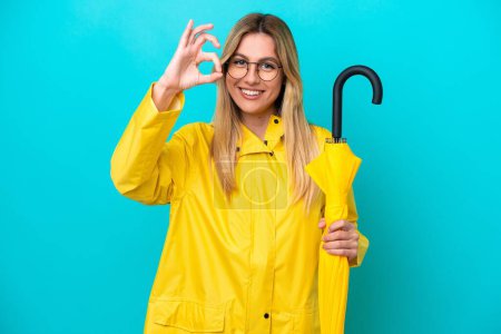 Young Uruguayan woman with rainproof coat and umbrella isolated on blue background showing ok sign with fingers