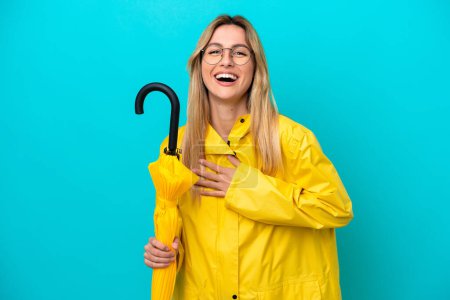 Young Uruguayan woman with rainproof coat and umbrella isolated on blue background smiling a lot