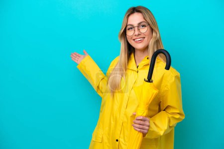 Young Uruguayan woman with rainproof coat and umbrella isolated on blue background extending hands to the side for inviting to come