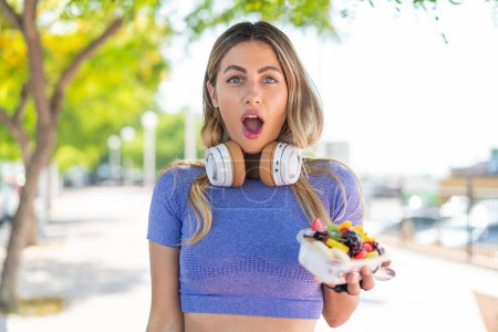 Téléchargez les photos : Jeune jolie femme sportive tenant un bol de fruits à l'extérieur avec une expression faciale surprise et choquée - en image libre de droit