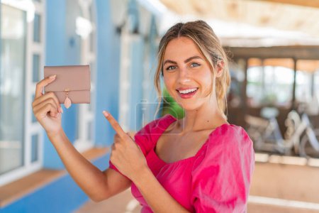 Photo for Young blonde woman holding a wallet at outdoors and pointing it - Royalty Free Image