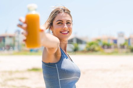 Téléchargez les photos : Jeune jolie femme sportive tenant un jus d'orange à l'extérieur avec une expression heureuse - en image libre de droit