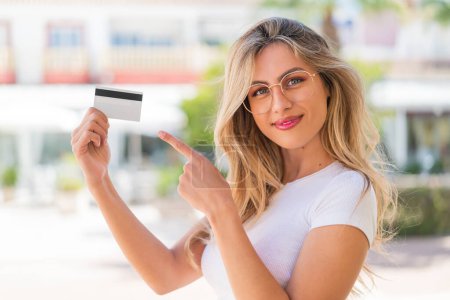 Téléchargez les photos : Jolie femme uruguayenne blonde tenant une carte de crédit à l'extérieur et la pointant du doigt - en image libre de droit
