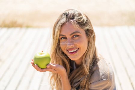 Téléchargez les photos : Jeune jolie femme uruguayenne blonde en plein air tenant une pomme avec une expression heureuse - en image libre de droit