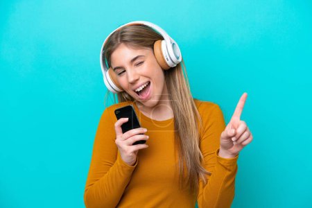 Téléchargez les photos : Jeune femme caucasienne isolée sur fond bleu écoutant de la musique avec un mobile et chantant - en image libre de droit