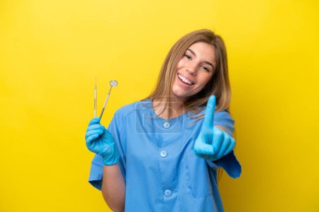 Photo for Dentist caucasian woman holding tools isolated on yellow background showing and lifting a finger - Royalty Free Image