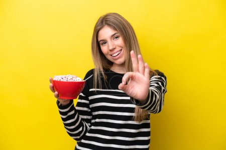 Téléchargez les photos : Jeune femme caucasienne tenant un bol de céréales isolé sur fond jaune montrant ok signe avec les doigts - en image libre de droit