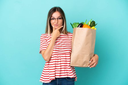 Foto de Mujer joven sosteniendo una bolsa de la compra de comestibles aislado en el pensamiento de fondo azul - Imagen libre de derechos