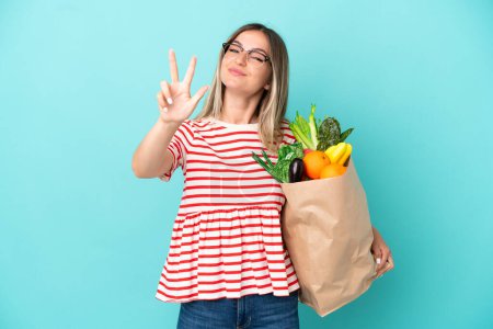 Téléchargez les photos : Jeune femme tenant un sac d'épicerie isolé sur fond bleu heureux et comptant trois avec les doigts - en image libre de droit