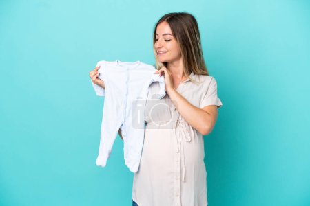 Téléchargez les photos : Jeune femme roumaine isolée sur fond bleu enceinte et tenant des vêtements de bébé - en image libre de droit