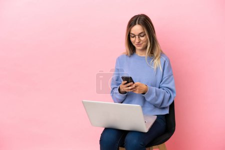 Téléchargez les photos : Jeune femme assise sur une chaise avec ordinateur portable sur fond rose isolé envoyer un message avec le mobile - en image libre de droit