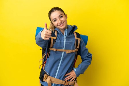 Téléchargez les photos : Jeune alpiniste femme avec un grand sac à dos sur fond jaune isolé avec les pouces vers le haut parce que quelque chose de bon est arrivé - en image libre de droit