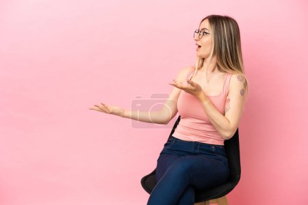 Téléchargez les photos : Jeune femme assise sur une chaise sur fond rose isolé avec expression faciale surprise - en image libre de droit