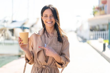 Foto de Mujer joven al aire libre sosteniendo un café para llevar al aire libre y señalándolo - Imagen libre de derechos