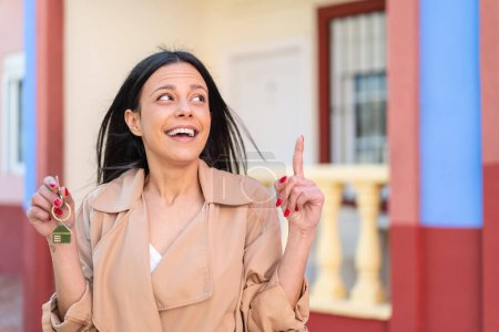 Téléchargez les photos : Jeune femme à l'extérieur tenant les clés de la maison à l'extérieur dans l'intention de réaliser la solution tout en levant le doigt vers le haut - en image libre de droit