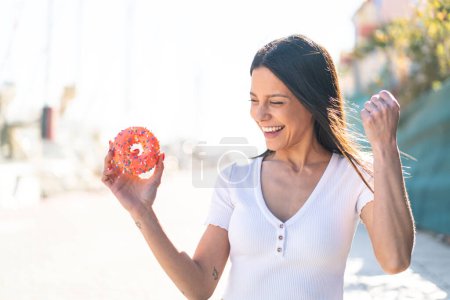 Téléchargez les photos : Jeune femme en plein air tenant un beignet en plein air célébrant une victoire - en image libre de droit