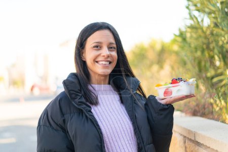 Téléchargez les photos : Jeune femme à l'extérieur tenant un bol de fruits à l'extérieur souriant beaucoup - en image libre de droit