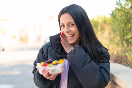 Téléchargez les photos : Jeune femme à l'extérieur tenant un bol de fruits à l'extérieur chuchotant quelque chose - en image libre de droit