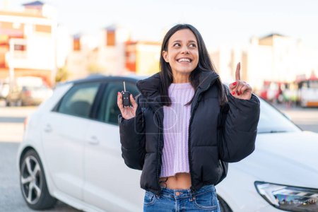 Téléchargez les photos : Jeune femme à l'extérieur tenant les clés de voiture à l'extérieur dans l'intention de réaliser la solution tout en levant le doigt vers le haut - en image libre de droit