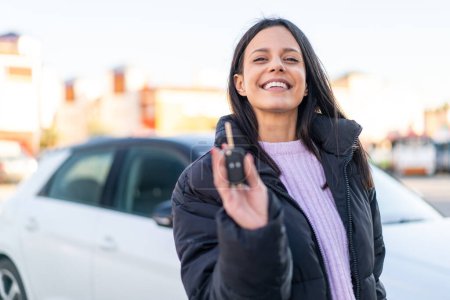 Téléchargez les photos : Jeune femme à l'extérieur tenant des clés de voiture à l'extérieur avec une expression heureuse - en image libre de droit