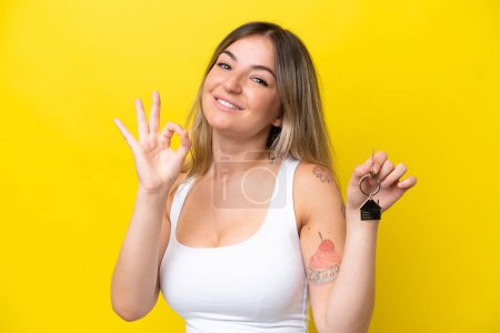 Téléchargez les photos : Jeune femme roumaine tenant les clés de la maison isolées sur fond jaune montrant ok signe avec les doigts - en image libre de droit
